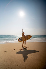 A young woman walks along the beach with a surfboard on the background of the ocean and the sun