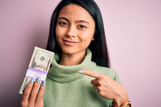 Young beautiful chinese woman holding dollars standing over isolated pink background very happy pointing with hand and finger