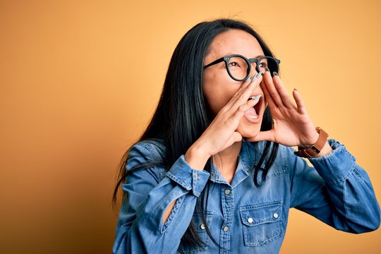 Young beautiful chinese woman wearing casual denim shirt over isolated yellow background Shouting angry out loud with hands over mouth