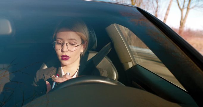 Pretty Caucasian young blonde businesswoman painting lips with red lipstick while looking in the mirror and sitting in the expensive car.