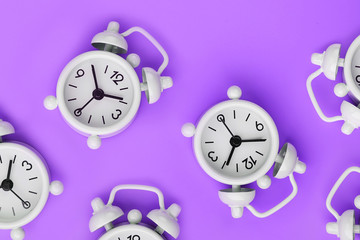 A pattern of many white classic alarm clocks in the form of a pattern on a purple background. Top view with a copy of the space, flat lay.