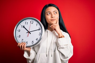 Young beautiful chinese woman holding big clock standing over isolated red background serious face...