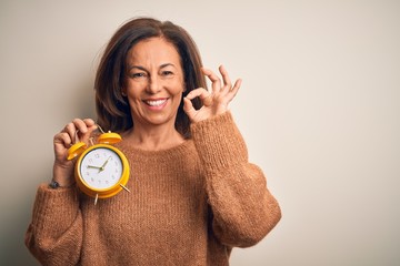 Middle age brunette woman holding clasic alarm clock over isolated background smiling positive doing ok sign with hand and fingers. Successful expression.