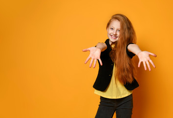 Ginger teenager in black jacket and pants, yellow t-shirt. She smiling, reaching for you, posing on orange background. Close up