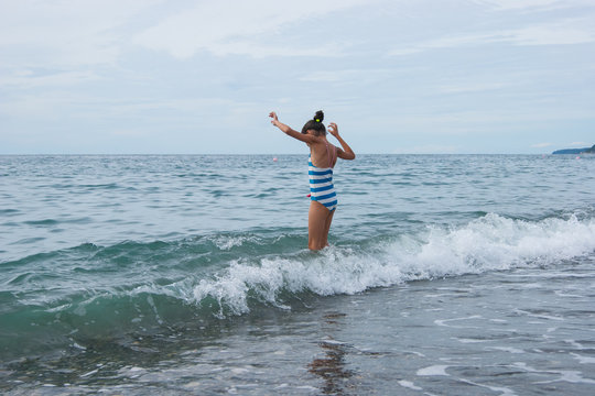 A girl in a blue-and-white stripe swimsuit enters the sea. Summer, waves, sea, beach, vacation, happiness.