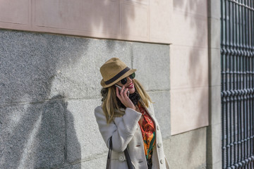 Blonde girl with coat and black glasses smiles while talking on her mobile phone in a city street