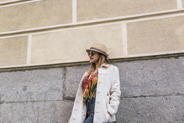 Fototapeta na wymiar Cute and attractive blonde woman with white coat and colorful clothes leaning on a wall