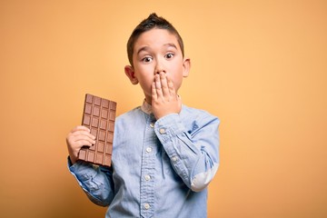 Young little boy kid eating sweet chocolate bar for dessert over isolated yellow background cover...