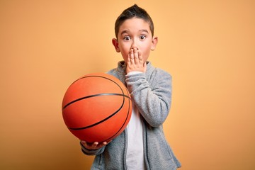 Young little boy kid playing with basketball game ball over isolated yellow background cover mouth with hand shocked with shame for mistake, expression of fear, scared in silence, secret concept