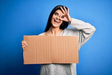 Young beautiful brunette activist woman holding blank cardboard banner protesting with happy face smiling doing ok sign with hand on eye looking through fingers