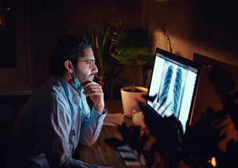 Young pensive doctor is skeptical about x-ray image on the monitor at dark office.