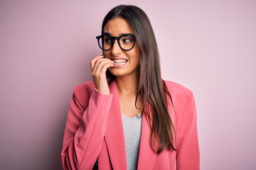 Young beautiful brunette businesswoman wearing jacket and glasses over pink background looking stressed and nervous with hands on mouth biting nails. Anxiety problem.