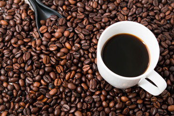 Roasted coffee beans scattered from white cup on dark wooden surface.