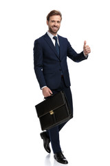 happy young businessman holding suitcase and making thumbs up sign