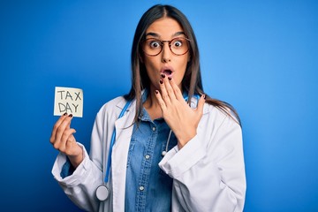 Young doctor woman wearing stethoscope holding paper with tax day message cover mouth with hand...