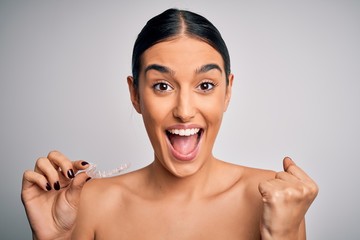 Young beautiful brunette woman holding dental aligner orthodontic to teeth corretion screaming proud and celebrating victory and success very excited, cheering emotion