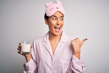 Young beautiful woman wearing pajama and sleep mask drinking glass of healthy milk pointing and showing with thumb up to the side with happy face smiling