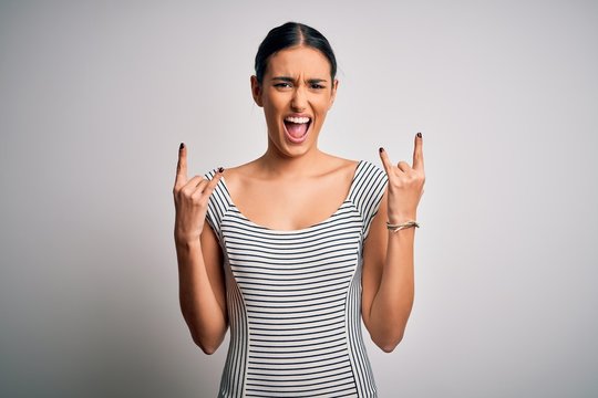 Young beautiful brunette woman wearing casual striped dress over isolated white background shouting with crazy expression doing rock symbol with hands up. Music star. Heavy concept.