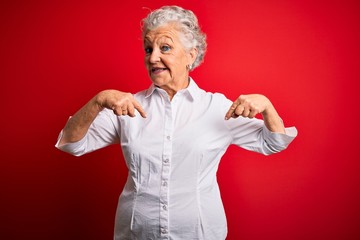 Senior beautiful woman wearing elegant shirt standing over isolated red background looking confident with smile on face, pointing oneself with fingers proud and happy.