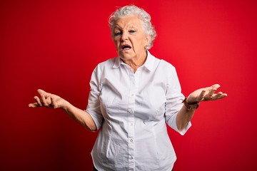 Senior beautiful woman wearing elegant shirt standing over isolated red background clueless and confused with open arms, no idea concept.