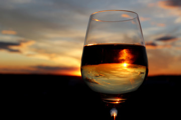 Glass with white wine on beautiful sunset background, sun and sky are reflected in alcohol drink. Concept of celebration, evening party at resort, romantic dinner outdoors