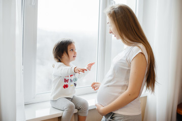 A pregnant mother is standing near the window with her little daughter