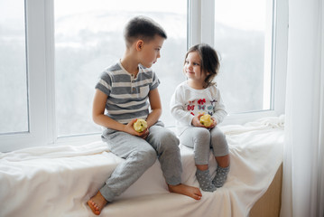 Obraz na płótnie Canvas Brother and sister are sitting on the windowsill playing and eating apples. Happiness