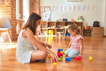 Beautiful teacher and blond toddler girl building tower using plastic blocks at kindergarten