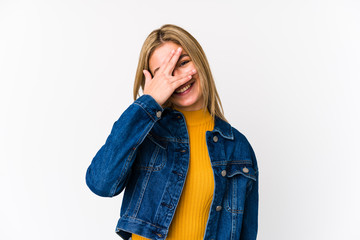 Young blonde caucasian woman isolated blink at the camera through fingers, embarrassed covering face.