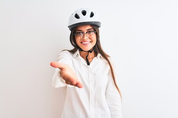Beautiful businesswoman wearing glasses and bike helmet over isolated white background smiling friendly offering handshake as greeting and welcoming. Successful business.
