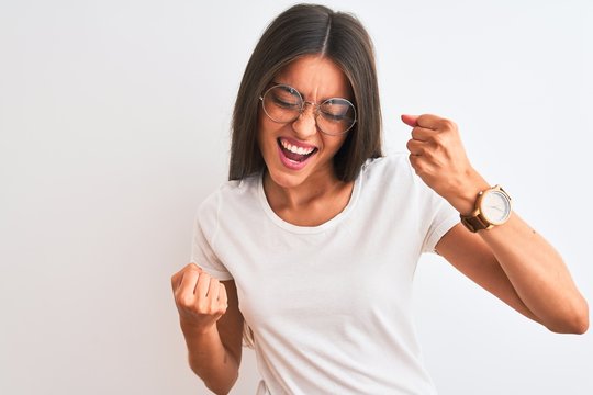Young beautiful woman wearing casual t-shirt and glasses over isolated white background very happy and excited doing winner gesture with arms raised, smiling and screaming for success. Celebration