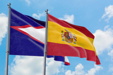 Spain and American Samoa flags waving in the wind against white cloudy blue sky together. Diplomacy concept, international relations.