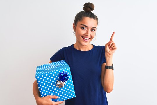 Young beautiful woman holding birthday gift standing over isolated white background very happy pointing with hand and finger to the side