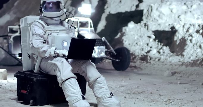 Caucasian man astronaut in the NASA armor costume sitting outside in the Moon valley and texting on the laptop computer.