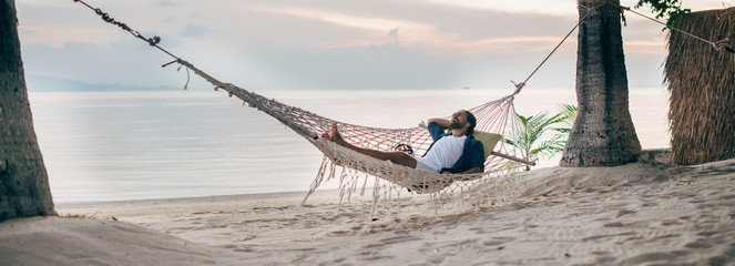 A man enjoys calm, lies in a hammock on the background of the ocean and sunset.