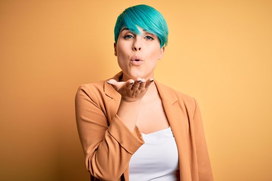 Young beautiful woman with blue fashion hair wearing casual jacket over yellow background looking at the camera blowing a kiss with hand on air being lovely and sexy. Love expression.