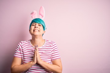 Young woman with fashion blue hair wearing easter rabbit ears over pink background begging and praying with hands together with hope expression on face very emotional and worried. Begging.