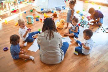 Beautiful teacher and group of toddlers sitting on the floor drawing using paper and pencil around lots of toys at kindergarten