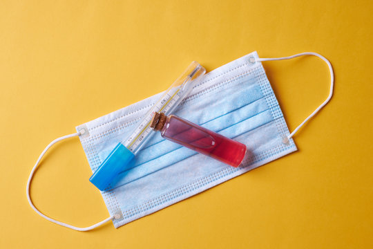 vial with red liquid over blue face mask thermometer on yellow background. coronavirus and infection protection concept - Powered by Adobe