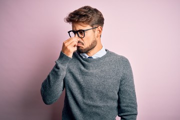 Young handsome man with beard wearing glasses and sweater standing over pink background smelling something stinky and disgusting, intolerable smell, holding breath with fingers on nose. Bad smell