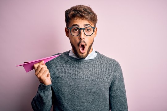 Young handsome man holding paper aircraft wearing glasses over isolated pink background scared in shock with a surprise face, afraid and excited with fear expression