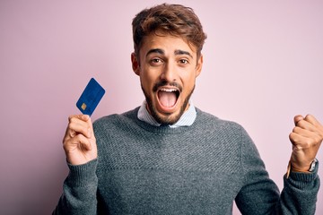 Young customer man with beard holding credit card for payment over pink background screaming proud and celebrating victory and success very excited, cheering emotion