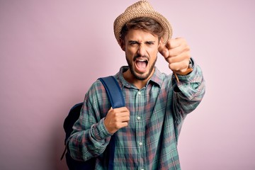 Young tourist man on vacation wearing backpack and summer hat over pink background annoyed and frustrated shouting with anger, crazy and yelling with raised hand, anger concept