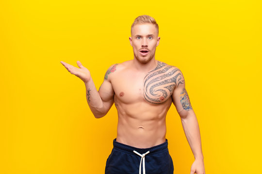 Young Strong Blonde Man Looking Surprised And Shocked, With Jaw Dropped Holding An Object With An Open Hand On The Side Against Yellow Wall