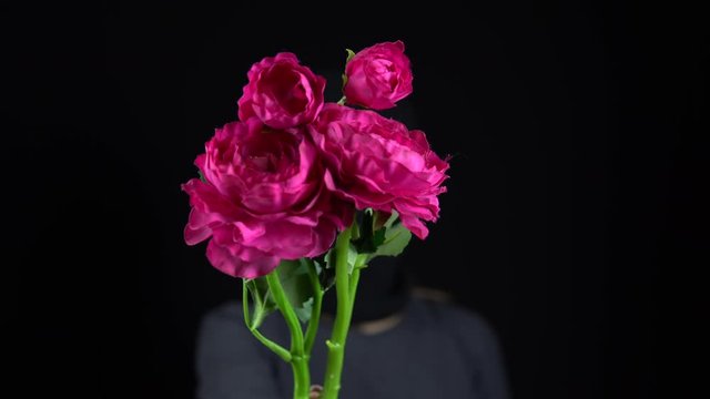 A woman in a balaclava mask is standing with flowers. The bandit holds out a bouquet of pink flowers to the camera. On a black background.