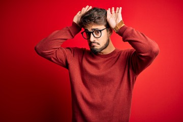 Young handsome man with beard wearing glasses and sweater standing over red background Doing bunny ears gesture with hands palms looking cynical and skeptical. Easter rabbit concept.