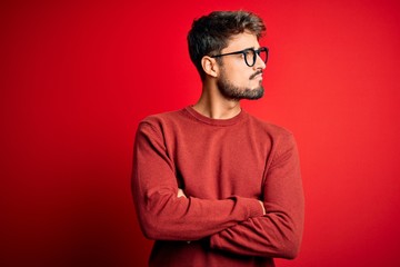 Fototapeta na wymiar Young handsome man with beard wearing glasses and sweater standing over red background looking to the side with arms crossed convinced and confident