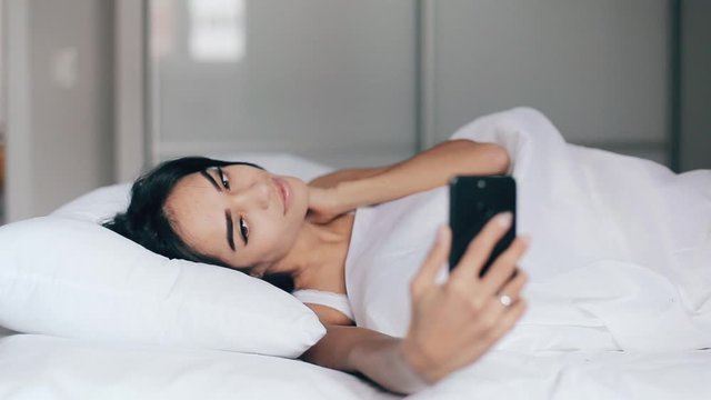 Pretty young girl in a bed takes selfies using a smartphone