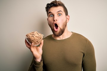 Young blond man with beard and blue eyes holding bowl with cornflakes cereals scared in shock with a surprise face, afraid and excited with fear expression