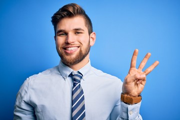 Young blond businessman with beard and blue eyes wearing elegant shirt and tie standing showing and pointing up with fingers number three while smiling confident and happy.